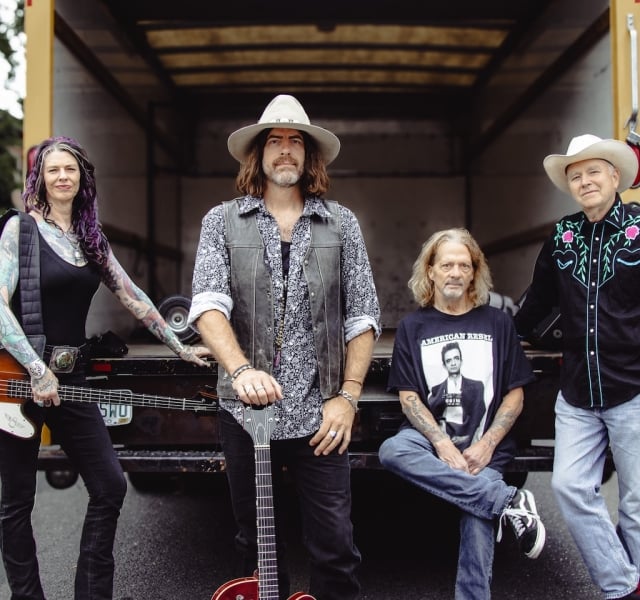 Four musicians posing in a truck bed: two women on the ends and two men center and right, with guitars and casual outfits.