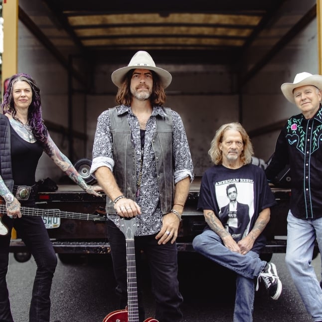 Four musicians posing in a truck bed: two women on the ends and two men center and right, with guitars and casual outfits.