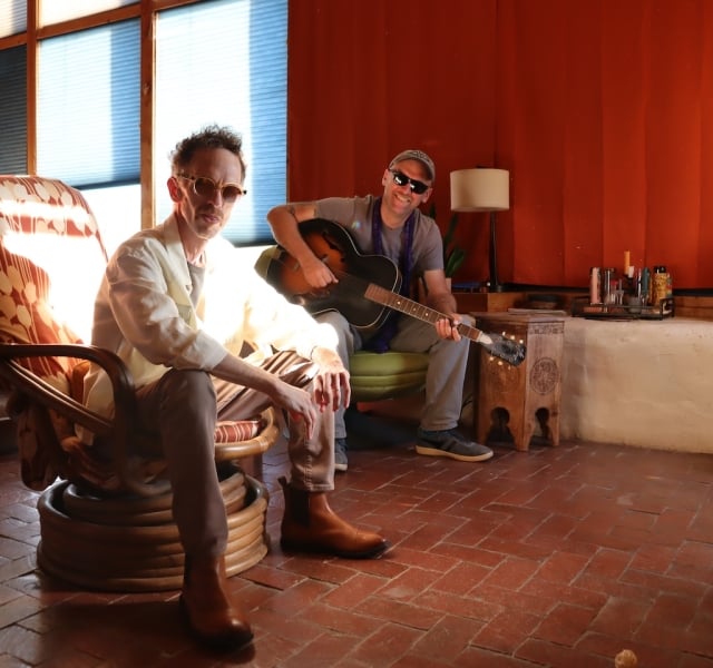 Two men in a sunlit, cozy room; one sits in a patterned chair while the other strums an acoustic guitar.