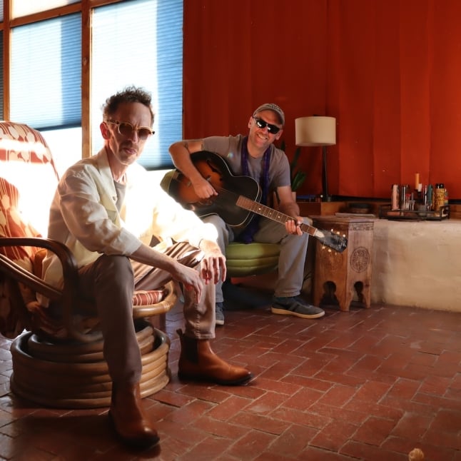 Two men in a sunlit, cozy room; one sits in a patterned chair while the other strums an acoustic guitar.