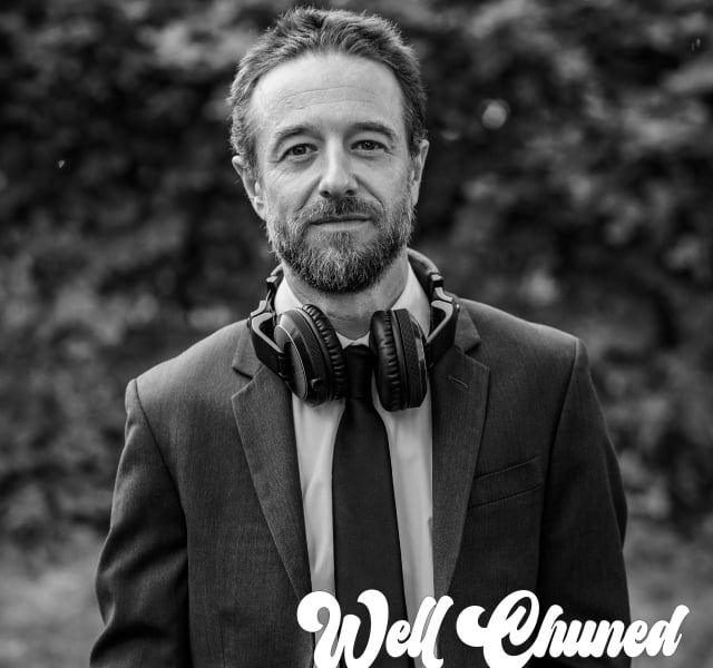 Close-up portrait of a bearded man in a suit with headphones around his neck, outdoors with a blurred background.