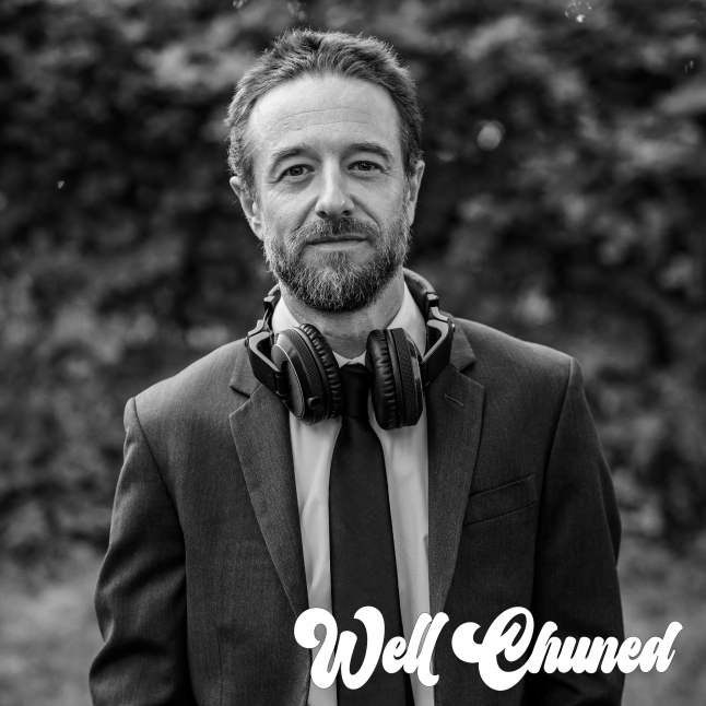 Close-up portrait of a bearded man in a suit with headphones around his neck, outdoors with a blurred background.