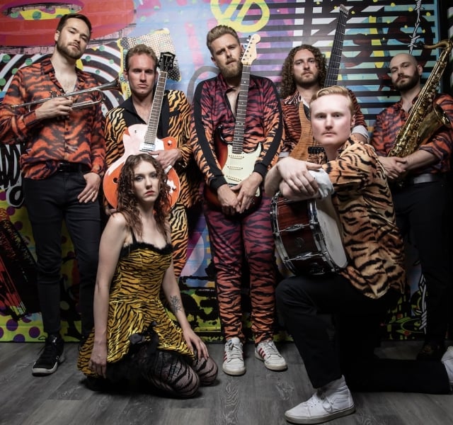 Seven musicians in tiger-stripe outfits pose in a colorful graffiti studio, holding brass, guitar, bass, drums, and a saxophone. A woman in a tiger‑print dress kneels in front.