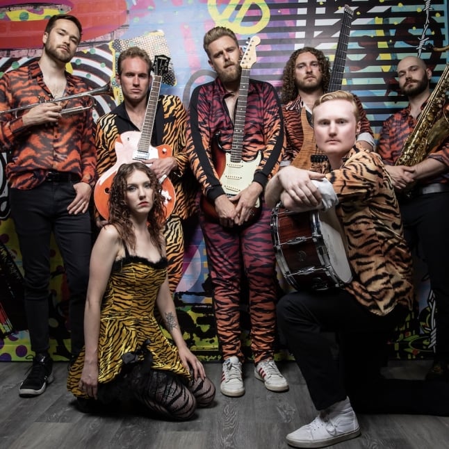 Seven musicians in tiger-stripe outfits pose in a colorful graffiti studio, holding brass, guitar, bass, drums, and a saxophone. A woman in a tiger‑print dress kneels in front.