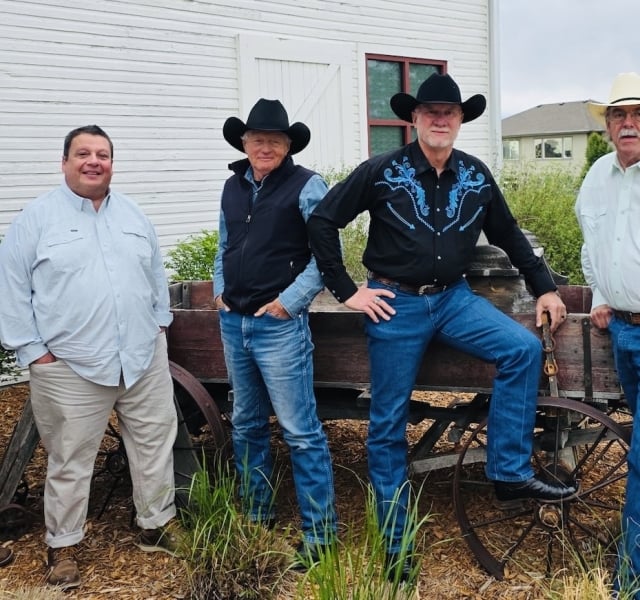 Five men in cowboy hats pose beside an old wooden wagon in a yard of a white building setting.