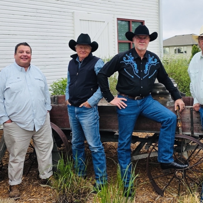 Five men in cowboy hats pose beside an old wooden wagon in a yard of a white building setting.