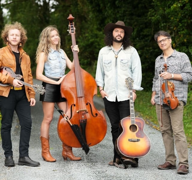 Four musicians posing outdoors on a gravel path with their instruments: mandolin, upright bass, acoustic guitar, and violin.