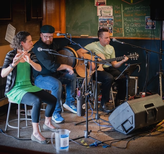Three musicians performing live on a small stage: a flutist, a percussionist, and a guitarist, with a chalkboard and posters behind them.