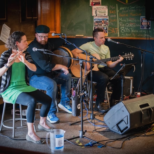 Three musicians performing live on a small stage: a flutist, a percussionist, and a guitarist, with a chalkboard and posters behind them.