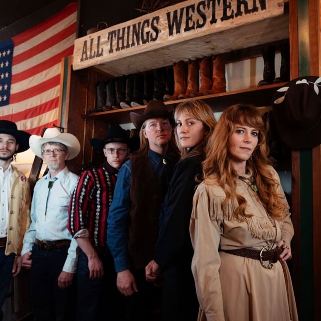 Six adults in cowboy hats and fringed jackets pose inside a western-themed shop, boots on display and a large American flag in the background.