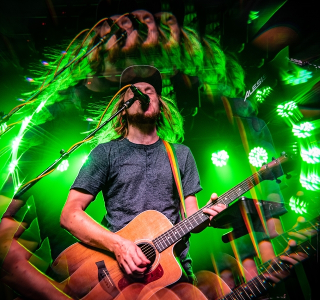 Male guitarist with long hair and a beard sings into a microphone while playing an acoustic guitar under vivid green stage lights on a live stage.