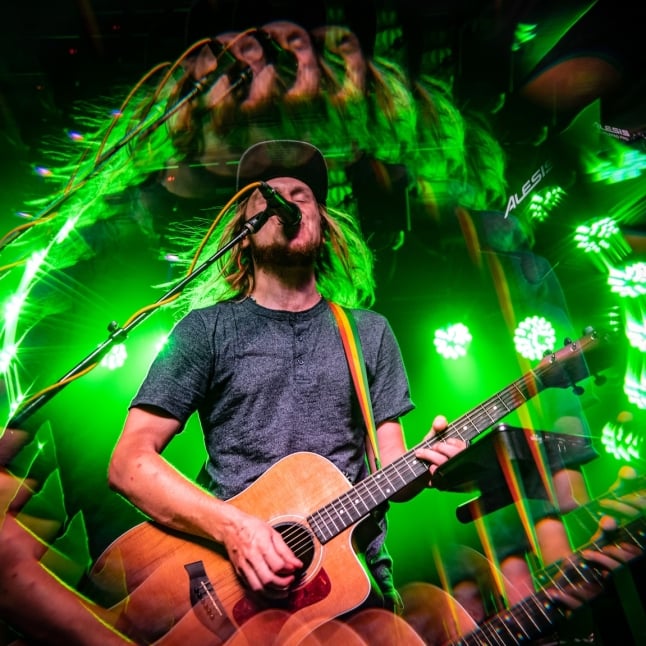 Male guitarist with long hair and a beard sings into a microphone while playing an acoustic guitar under vivid green stage lights on a live stage.