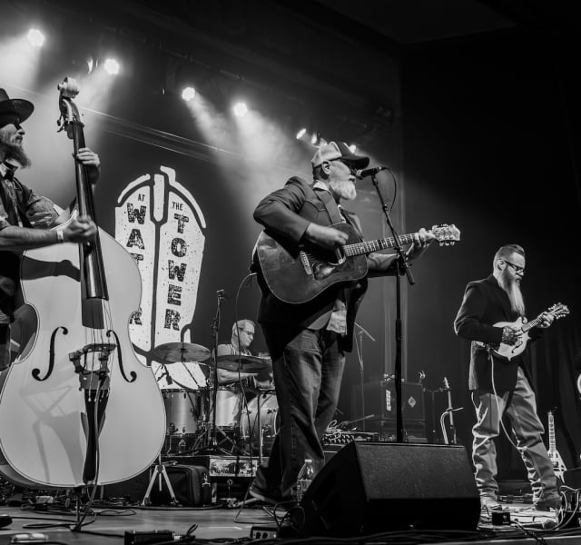 Black-and-white photo of a five-member band performing on a crowded stage with guitars, bass, drums, and a vocalist at center under stage lights while a drummer plays in the back.