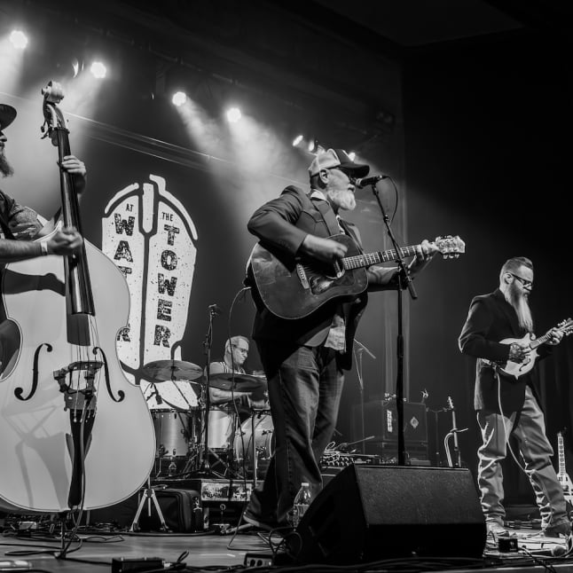 Black-and-white photo of a five-member band performing on a crowded stage with guitars, bass, drums, and a vocalist at center under stage lights while a drummer plays in the back.