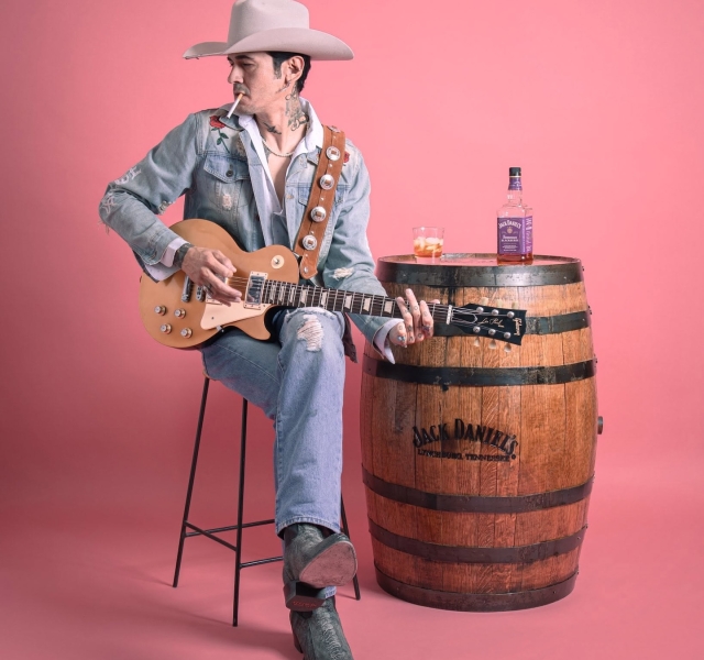 Musician in a beige cowboy hat and denim jacket plays a guitar, cigarette in mouth, seated beside a whiskey barrel with a bottle and glass on top.