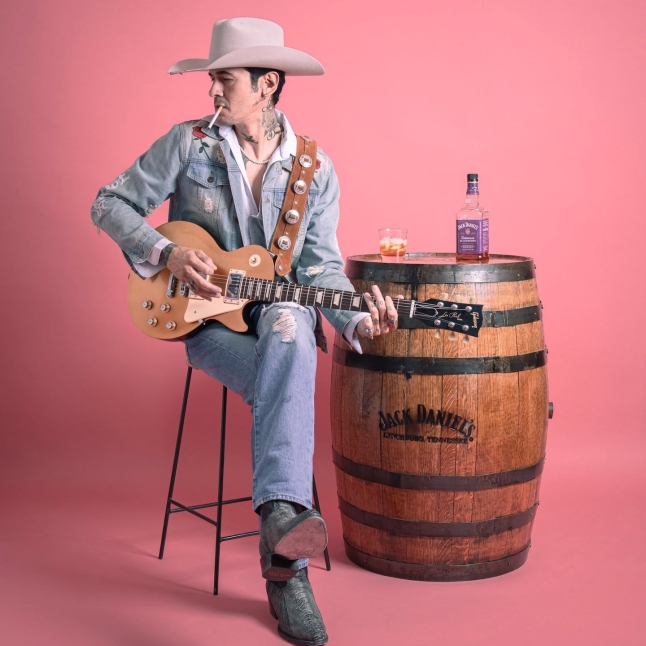 Musician in a beige cowboy hat and denim jacket plays a guitar, cigarette in mouth, seated beside a whiskey barrel with a bottle and glass on top.