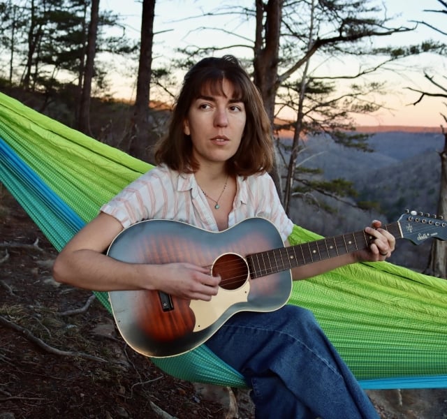 Person sitting in a bright green hammock outdoors, playing an acoustic guitar at sunset in a wooded setting.