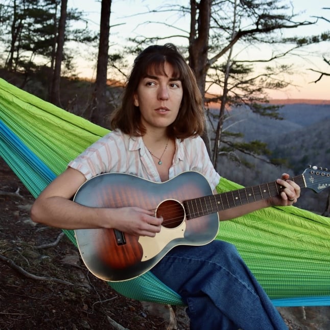 Person sitting in a bright green hammock outdoors, playing an acoustic guitar at sunset in a wooded setting.