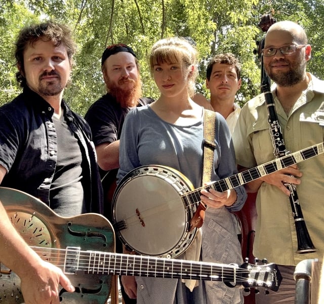 Group of five musicians posing outdoors under leafy trees, holding a guitar, banjo, and a clarinet/sax-style instrument. Intimate, casual band photo.