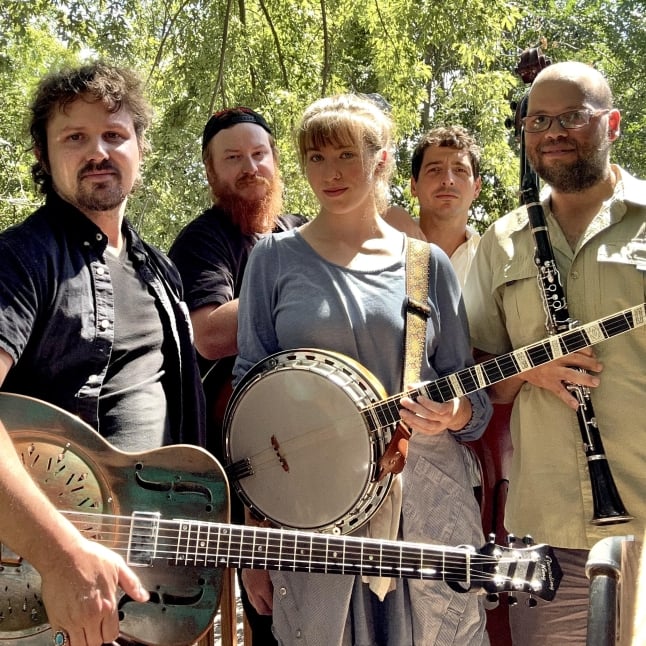 Group of five musicians posing outdoors under leafy trees, holding a guitar, banjo, and a clarinet/sax-style instrument. Intimate, casual band photo.