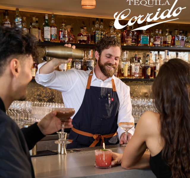 Bartender with a beard pours a cocktail from a shaker for two customers at a bar, Tequila Corrido logo in the corner behind them.