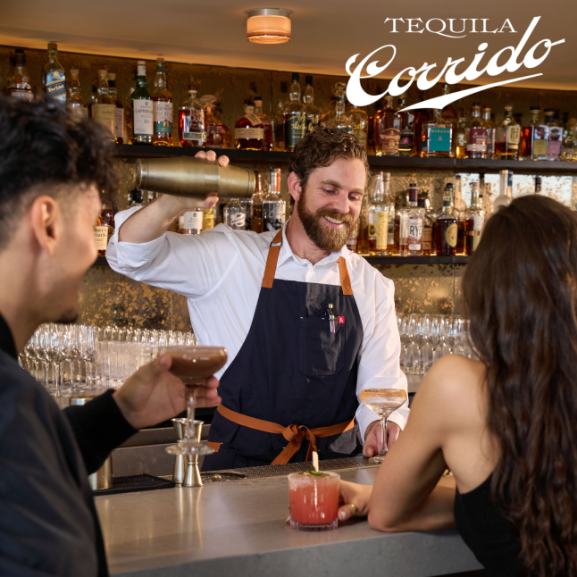 Bartender with a beard pours a cocktail from a shaker for two customers at a bar, Tequila Corrido logo in the corner behind them.