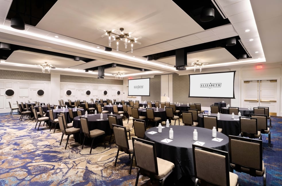 Conference banquet setup in a large room with round tables, black tablecloths, water bottles, and chairs facing two projection screens showing a hotel logo.